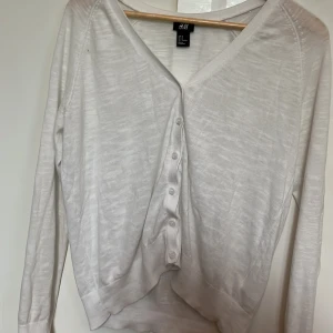 Vit kofta från H&M med knappar - Vit tunn kofta från H&M i storlek M. Koftan har v-ringning, långa ärmar och stängs med små vita knappar framtill. Perfekt att slänga över en topp för en clean och enkel look. Materialet känns lätt och luftigt.