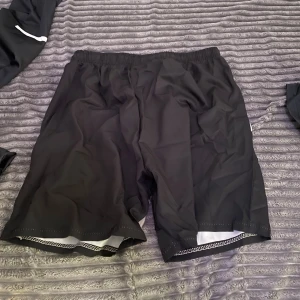 Svarta Nike shorts med snörning - Nike shorts i svart med bred elastisk midja och vit snörning. De har en grå panel med Nike-logga på ena benet och är gjorda i lätt polyester som passar perfekt för träning eller sport. Snygg och sportig look med normal passform.
