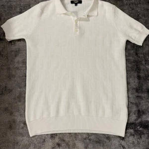 Vit Fendi piké  - Säljer en stilren vit piké från Fendi med diskret strukturmönster i tyget. Klassisk pikékrage och knappar framtill. Pikén är kortärmad och har ribbade kanter nedtill och vid ärmslut. Perfekt för dig som gillar exklusiva och cleana plagg.