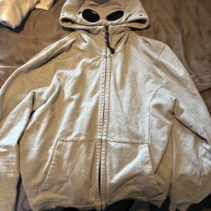 Cp company - Säljer en grå hoodie från C.P. Company i storlek XL. Tröjan har hel dragkedja, stora fickor framtill och en unik huva med inbyggda glasögon. Perfekt för dig som vill sticka ut med en streetwear-look.
