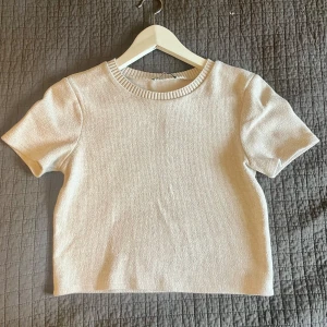 Beige stickad topp Zara - En stilren beige stickad topp från Zara med korta ärmar och ribbad rund halsringning. Toppen har en croppad passform och perfekt att matcha med jeans eller kjol för en clean look.