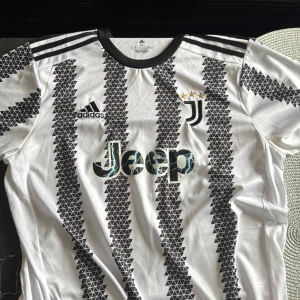 Juventus fotbollströja Adidas  - Juventus matchtröja från Adidas i storlek XL men passar mer som en L. Vit bas med svarta geometriska mönster och svarta detaljer på axlar och krage. Jeep-logga på bröstet, Juventus klubbmärke med tre stjärnor och Adidas-logga. Tillverkad i lätt polyester, perfekt för träning eller match även hemma använding. priset är ej hugget i sten