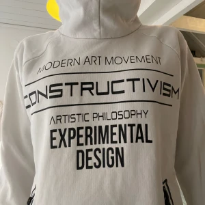 Vit hoodie med tryck från WOXO 720 - Vit hoodie från WOXO 720 med svart textprint över hela framsidan och nederkanten. Tröjan har huva, långa ärmar och dragkedjor vid fickorna. Perfekt för dig som gillar streetstyle och grafiska detaljer. Snygg och unik. I storlek 150 till ca 170. 