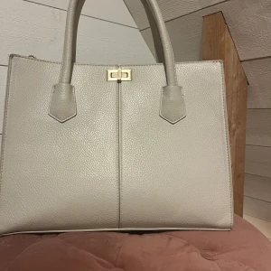 Beige handväska i skinn med gulddetalj - Stilren beige handväska i skinn med två rejäla handtag och snygg guldfärgad låsdetalj framtill. Väskan har en klassisk, fyrkantig form och är perfekt för dig som gillar minimalistisk design och neutrala färger.