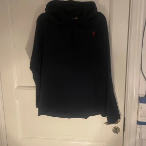 Svart hoodie från Polo Ralph Lauren - Svart hoodie från Polo Ralph Lauren med klassisk röd broderad logga på bröstet. Tröjan har huva med snörning och långa ärmar. Perfekt för en chill och stilren look. Tillverkad i mjuk bomull för extra komfort.