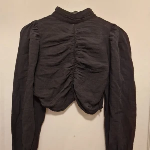 Svart blus Bianca x Nelly - Unik svart blus med hög krage och puffiga långärmar. Från Biancas x Nelly collection.  Croppad modell som passar perfekt till höga byxor eller kjol.