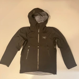 Svart vindjacka från Arc'teryx M - Säljer en svart vindjacka från Arc'teryx i storlek M. Jackan har huva, vattentäta dragkedjor och två snedställda fickor framtill. Perfekt för dig som gillar stilren och funktionell design. Materialet är syntet och jackan har lång ärm.