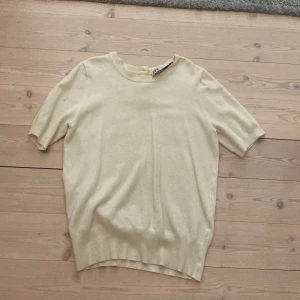 Beige kortärmad tröja från Zara - Snygg beige tröja från Zara med korta ärmar och rund hals. Tröjan har ribbad nederkant och tre dekorativa knappar bak i nacken. Perfekt till vår och sommar, enkel att matcha och supermjuk mot huden.