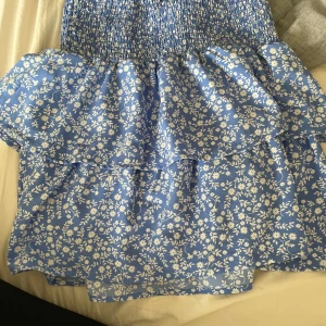 Blå blommig volangkjol från Seaside - Supersöt blå kortkjol från Seaside med vitt blommönster och volangdetaljer. Kjolen har smockad midja som sitter skönt och ger en snygg passform. Perfekt för sommarens alla äventyr!