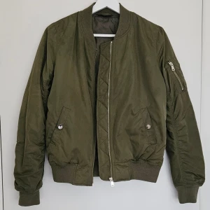 Militärgrön Bomberjacka  - Klassisk bomberjacka i militärgrön/khaki från TopShop. Modellen har fickor fram med tryckknappar samt en ärmficka med dragkedja och ribbade muddar i ärmslut, midja och kragen.       Väldigt fint skick, Strl S 