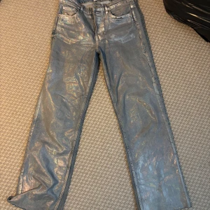 Glittriga silverfärgade jeansbyxor - Raka, lågmidjade jeans från zara i storlek 38. Jeansen är blå under men har en skimrande och metalliskt effekt i främst silver som reflekteras i ljuset till olika nyanser som blå, grön och guld. Detta ger en holografisk finish. Dem har fyra knappar framtill som man knäpper. Byxorna passar mig som bär 32-38 och kan sitta lite pösigare vid låren eller tight beroende på vilken storlek man har. Super snygga och coola.  SKICKAS SAMMA DAG SOM KÖP