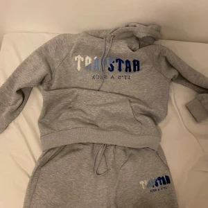 Grå hoodie och byxor från Trapstar med tryck - Säljer en grå hoodie och byxor från Trapstar med fet logga i vitt och blått framtill. Klassisk känguruficka, snörning i huvan och mjukt material. Perfekt för chill dagar och streetwear-vibe. Storlek L. Setet har inte används alls och är helt nytt! Kontakta mig vid behov av fler bilder!