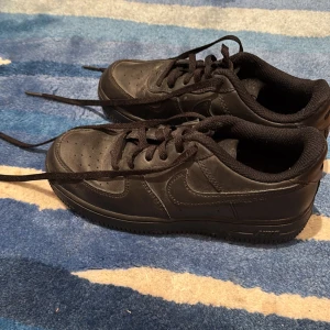 Svarta Nike Air Force 1 sneakers - Klassiska helsvarta Nike Air Force 1 sneakers i skinn med lågt skaft och rund tå. Skorna har svart snörning och diskret Nike-logga på sidan. Perfekta för en clean och stilren look. Pris kan diskuteras.