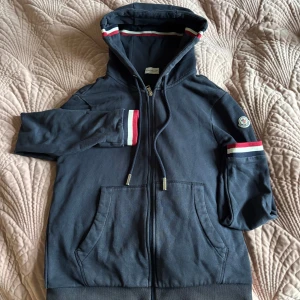 Marinblå Moncler hoodie med huva - Snygg marinblå hoodie från Moncler med huva och dragkedja. Tröjan har långa ärmar och coola ränder i rött, vitt och blått på muddar och insidan av huvan. Moncler-logga på vänster ärm och praktiska fickor framtill. Perfekt för chill dagar.