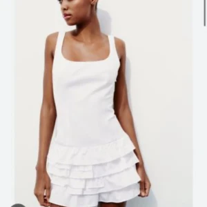 Vit kort klänning med volanger - Superfin vit kort klänning med breda axelband och fyrkantig urringning. Kjolen har flera lager volanger som ger en lekfull och trendig look. Perfekt för sommarens alla tillfällen. Sval och lätt i materialet.