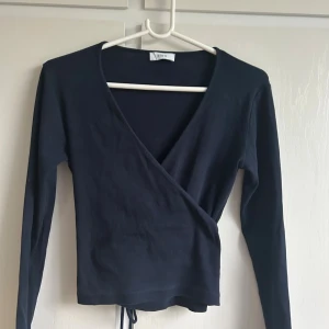 Svart omlottopp från Envii - Svart omlottopp från Envii med lång ärm och v-ringning. Toppen har knyt i midjan och är tillverkad i ett ribbat material som ger en snygg passform. Perfekt att styla med jeans 