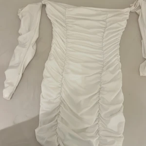Vit offshoulder kortklänning - Säljer en vit kortklänning med offshoulder och långa ärmar. Klänningen har snygga rynkade detaljer längs hela framsidan som ger en cool och trendig look. Perfekt för dig som gillar stilrena och moderna plagg med lite edge.