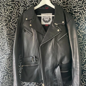 Svart skinnjacka Brando long S - Riktigt snygg svart mc jacka från Brando. Jackan har flera dragkedjor, bälte i midjan och detaljer i silver. Gjord i äkta skinn med bred krage och asymmetrisk dragkedja framtill. Använd två gånger. Har ett litet märka som knappt syns. Finns på bild!