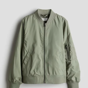 Grön bomberjacka från H&M - Snygg ljusgrön bomberjacka från H&M med klassisk ribbad krage och muddar. Jackan är aldrig använd och har dragkedja framtill, fickor på sidorna och en extra ficka med dragkedja på ärmen. Perfekt för dig som gillar en chill och stilren look. Säljer pga är lite för stor för mig som är relativt kort.