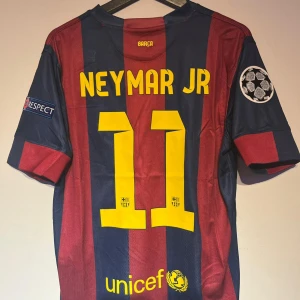 Barcelona Neymar JR matchtröja - Säljer en FC Barcelona matchtröja med Neymar JR och nummer 11 på ryggen. Tröjan har breda marinblå och vinröda ränder, gula detaljer och Champions League-märke på ärmen. Broderat klubbmärke och Qatar Airways-tryck framtill. Tillverkad i lätt polyester.