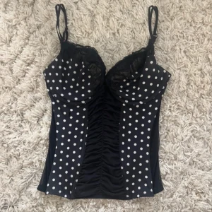 Prickig korsett topp - Snygg svart korsett med vita prickar och tunna axelband. Kuporna har spetsdetaljer och framsidan är rynkad för en cool effekt. Stängs med hyskor i ryggen. Finns ett litet hål på framsidan men inget man märker av! 