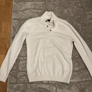 Vit polotröja från Bondelid med knappar - Snygg vit polotröja från Bondelid i storlek S. Tröjan har hög krage med tre svarta knappar framtill och diskret broderad logga. Mjuk stickad kvalitet som känns skön mot huden. Perfekt för dig som gillar stilrena och klassiska plagg.