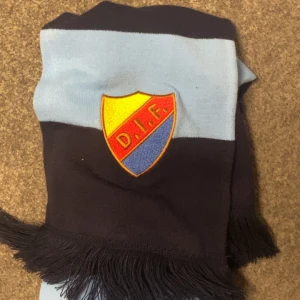 Blå DIF halsduk med fransar - Säljer en snygg halsduk från Djurgårdens IF med broderat klubbmärke. Halsduken har breda marinblå och ljusblå ränder samt svarta fransar i ändarna. Perfekt för att visa ditt stöd på matchen eller hänga med polarna.