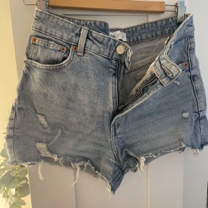 Blå jeansshorts med rå kant från Zara - Snygga blå jeansshorts från Zara i klassisk femficksmodell. De har medelhög midja, slitna detaljer och rå, fransig kant nedtill för en cool vibe. Materialet är robust denim med dragkedja och knapp framtill. Perfekta för sommarens alla äventyr.