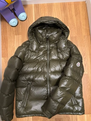 Mörkgrön pufferjacka från Moncler - Säljer en mörkgrön pufferjacka från Moncler med huva och dragkedja framtill. Jackan har ett glansigt yttermaterial, Moncler-logga på ärmen och praktiska fickor med dragkedja. Perfekt för kalla dagar och har en riktigt schysst streetstil.