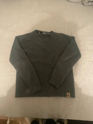Mörkgrå stickad tröja Calvin Klein - Mysig mörkgrå stickad tröja från Calvin Klein Jeans med rund hals och ribbade muddar. Tröjan har en enkel design med CK-logga på nederkanten och långa ärmar. Perfekt för kyliga dagar och enkel att matcha med jeans eller kjol.