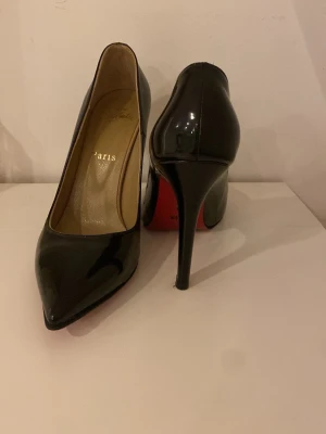 Svarta pumps  - Säljer ett par ikoniska svarta pumps  med glansig finish och klassisk röd sula. Skorna har spetsig tå och hög klack, tillverkade i skinn. Perfekta för dig som vill ha en elegant och tidlös look. Passar även 38