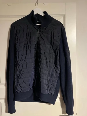 Mörkblå quiltad jacka från J.Lindeberg - Snygg mörkblå jacka från J.Lindeberg med quiltad framsida och stickade ärmar och rygg. Jackan har dragkedja framtill, högt krage och diskret logga på ärmen. Perfekt för dig som gillar stilren design och vill ha något som sticker ut lite.