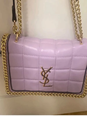 Ljuslila quiltad axelväska från YSL - Säljer en ljuslila quiltad axelväska från YSL med guldfärgade kedjor och detaljer. Väskan har ett stort YSL-emblem framtill och en praktisk dragkedja på baksidan. Tillverkad i skinn och har en lyxig känsla med snyggt rutmönster.