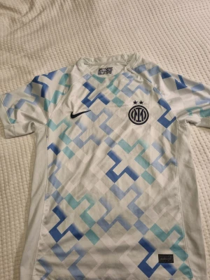 Inter Milan Dri-Fit t-shirt Nike - Säljer en Inter Milan t-shirt från Nike med Dri-Fit teknologi. Tröjan är vit med geometriskt mönster i blå och turkosa toner, kortärmad och har klubbens logga med två stjärnor på bröstet. 
