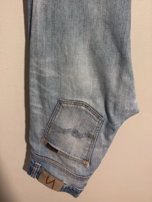Ljusblå jeans från Nudie Jeans - Säljer ett par ljusblå jeans från Nudie Jeans med klassisk femficksdesign och slitna detaljer. Jeansen har Nudie-loggan på bakfickan och patch i midjan. Tillverkade i mjukt denimtyg som ger en avslappnad vibe Modell: GRIM TIM. W:30 L:32