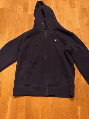 Marinblå Zip Hoodie Polo Ralph Lauren - Snygg blå hoodie från Polo Ralph Lauren med dragkedja och klassisk logga broderad på bröstet. Tröjan har huva med snörning och två stora fickor framtill. Perfekt för en clean och stilren look.                                           