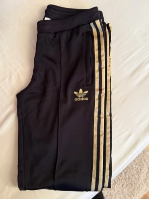 Nya Svarta Adidas trackpants med guldiga ränder - NYA. Svarta Adidas trackpants med guldiga ränder längs sidorna och broderad Adidas-logga på låret. Byxorna har ribbad midja, dragkedja vid benslut och är gjorda i mjukt syntetmaterial. Perfekta för chill eller träning, med klassisk streetstil. Strl 38