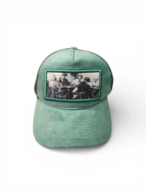 Grön keps från Emilio Pendenza - Snygg grön keps från Emilio Pendenza med mesh-bakstycke och justerbar knäppning. Framsidan har en unik patch med svartvit bild på personer runt ett bord. Perfekt accessoar för en cool och avslappnad stil.