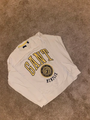 Vit sweatshirt från GANT med tryck - Vit sweatshirt från GANT med stort gult och blått tryck på bröstet och emblem. Rund hals med ribbad kant och gul detalj på insidan. Mjuk bomullskvalitet och långärmad modell, perfekt för chill dagar.