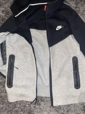 Nike tech svart och grå  - Nike vindjacka i svart och ljusgrått med huva och hel dragkedja. Två fickor med dragkedja framtill och vit piping längs huvan. Klassisk Nike-logga på bröstet. Perfekt för chill eller när du är på språng. Hör av er vid funderingar