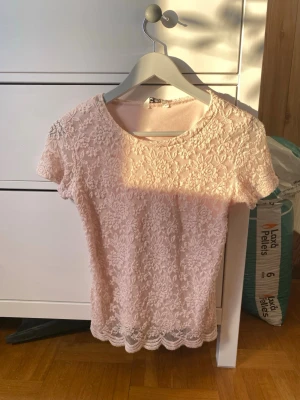 Ljusrosa spetsblus med blommönster - Säljer en ljusrosa blus med korta ärmar och rund hals. Blusen är täckt av ett detaljerat blommigt spetsmönster och har en vågig kant längst ner. Materialet är mjukt och luftigt, perfekt för vår och sommar. Supersöt och feminin stil! En liten noppa (se bild) som man inte tänker på, går såkert att klippa bort. Storlek 164 men passar XS