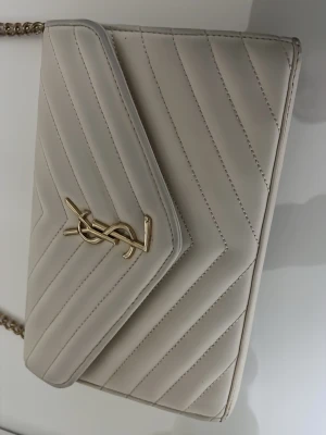 Beige quiltad handväska från Saint Laurent - Säljer en beige quiltad axelremsväska från Saint Laurent med ikonisk YSL-logga i guld på framsidan. Väskan är quiltad med snedställda sömmar och har kedjerem i guld. Insidan har flera fack och en dragkedja för smidig organisering. Perfekt för både vardag och fest! 