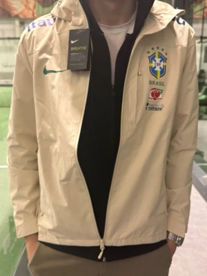 Nike Brasil vindjacka med huva - Säljer en beige Nike vindjacka med Brasil-logga och sponsordetaljer. Jackan har huva med dragsko, hel dragkedja och tryckta loggor på bröst och ärm. Perfekt för dig som gillar sportig stil och vill sticka ut med landslagsvibe.
