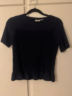 Mörkblå topp med volangkant Gina - Säljer en mörkblå topp med korta ärmar och rund hals. Framsidan har ett smockat parti som ger en cool textur och nederkanten är dekorerad med en söt volang. Perfekt att styla med jeans eller kjol för en chill look. Storlek 146-152