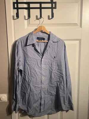 Randig blå skjorta från Ralph Lauren - Snygg skjorta från Ralph Lauren i blått och vitt med tunna ränder. Klassisk krage, långärmad och med knappar framtill. Tillverkad i 100% bomull med stretch för extra komfort. Ikonisk logga broderad på bröstet.