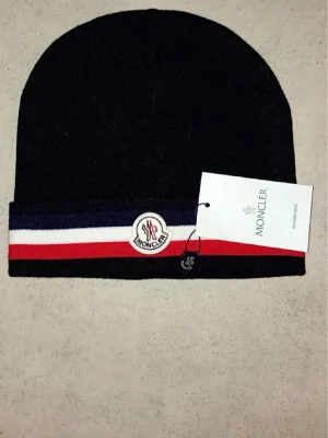 Svart Moncler mössa med ränder - Snygg svart mössa från Moncler med klassiska ränder i blått, vitt och rött längs kanten. Mössan har Moncler-logga framtill och är stickad i mjukt material som håller dig varm. Perfekt accessoar för att lyfta din look under kyliga dagar.