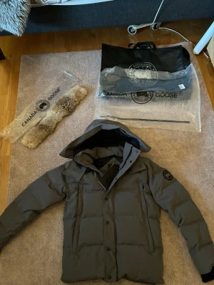 Mörkgrå Canada Goose xs  - Säljer en mörkgrå dunjacka från Canada Goose med huva och avtagbar pälsdetalj. Jackan har klassisk puffer-design, svarta knappar och logotyp på ärmen. Perfekt för kalla dagar och har en clean, modern look. Kommer med originalpåse och etiketter.