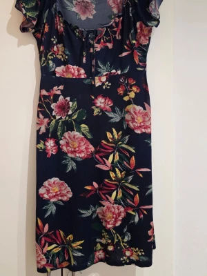 Blommig marinblå klänning Happy Holly - Säljer en marinblå kortklänning från Happy Holly i storlek 36/38. Klänningen har ett färgstarkt blommigt mönster i rosa, rött och grönt, med korta ärmar och knytning vid halsen. Materialet är mjuk och stretchig polyester med 5% elastan för extra komfort.