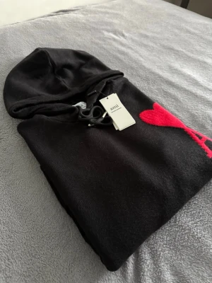 Ami Paris tröja  - Svart hoodie från Ami med huva och snörning. På bröstet finns ett stort rött hjärta med bokstaven A. Tröjan har ribbade muddar och en klassisk känguruficka framtill. Materialet är mjukt och stickat, perfekt för chill dagar.