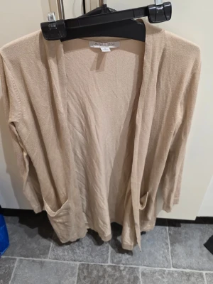 Beige kofta från Amisu i medium - Säljer en beige, långärmad kofta från Amisu i storlek medium. Koftan är stickad och har två praktiska fickor framtill. Materialet består av viskos, polyacryl och polyamid vilket gör den mjuk och skön. Perfekt att slänga på sig när det är lite kyligt ute.